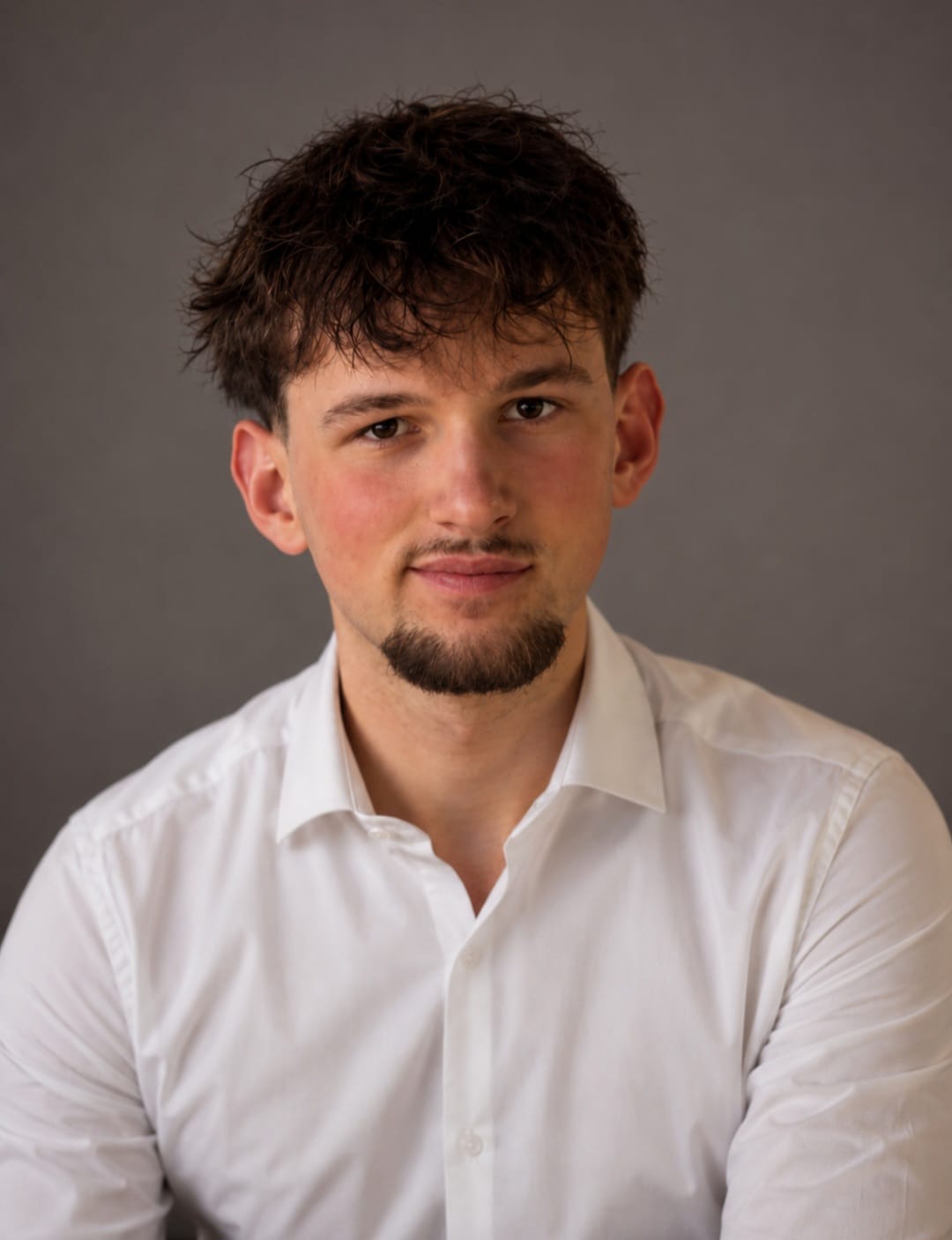 Young man with curly dark hair and goatee wearing a white button-up shirt against a gray background.
