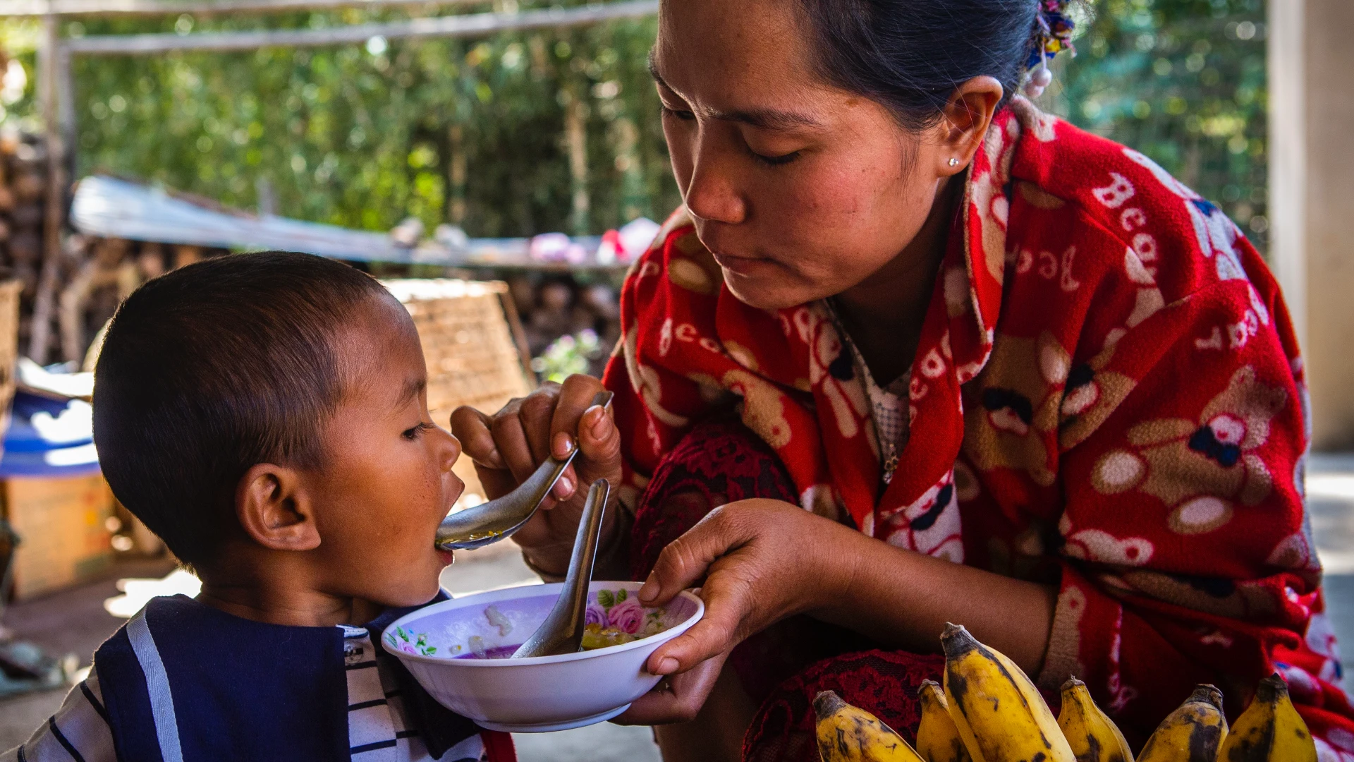 Photo of a mother feeding her daughter a nutritious meal cooked with ingredients grown in her backyard, Myanmar.