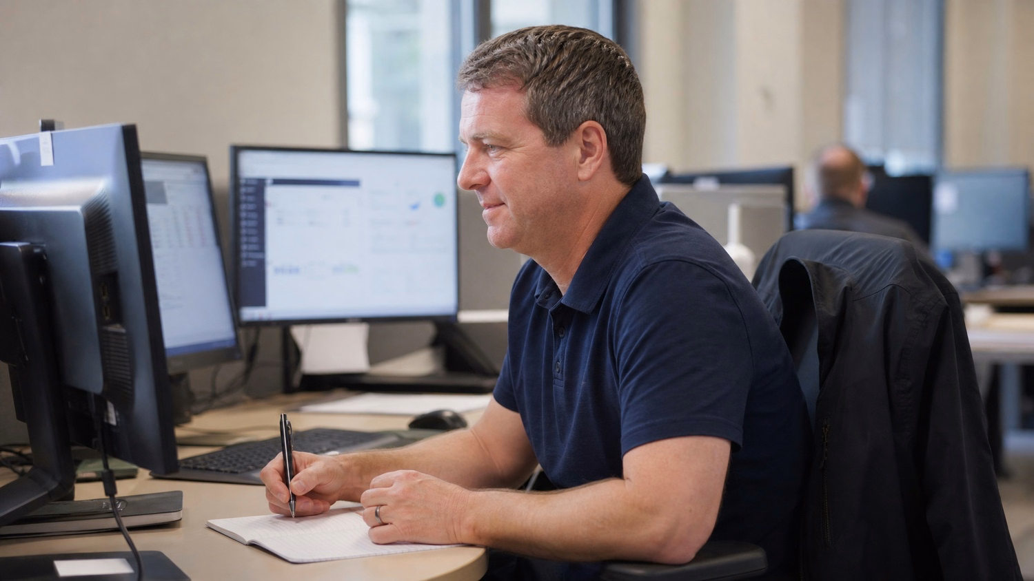 Man in polo shirt works on computer, notebook nearby