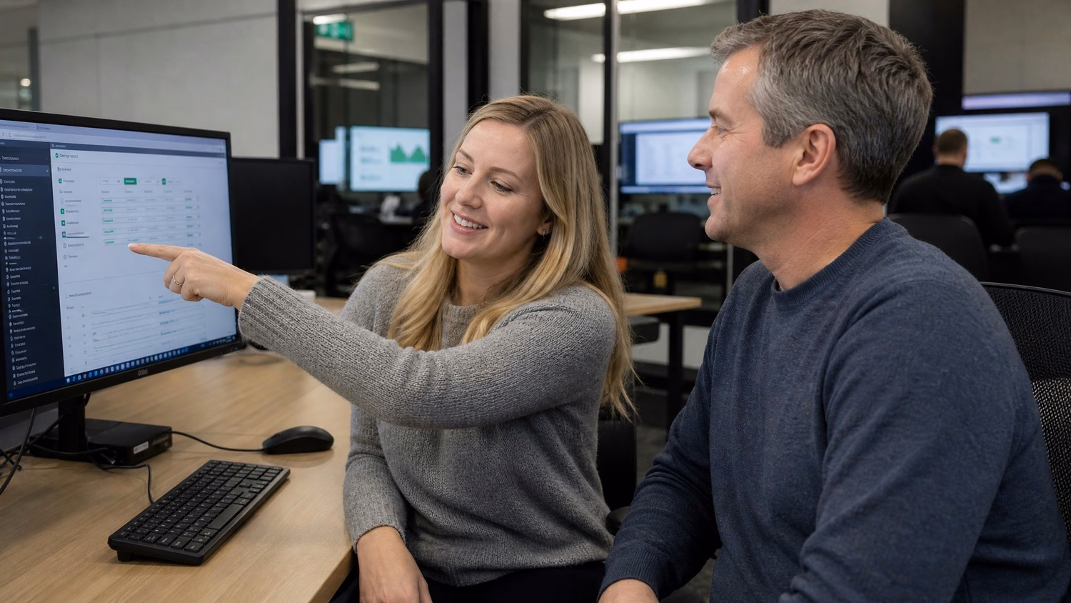 Woman points at monitor, colleague watches, shared desk 61 chars