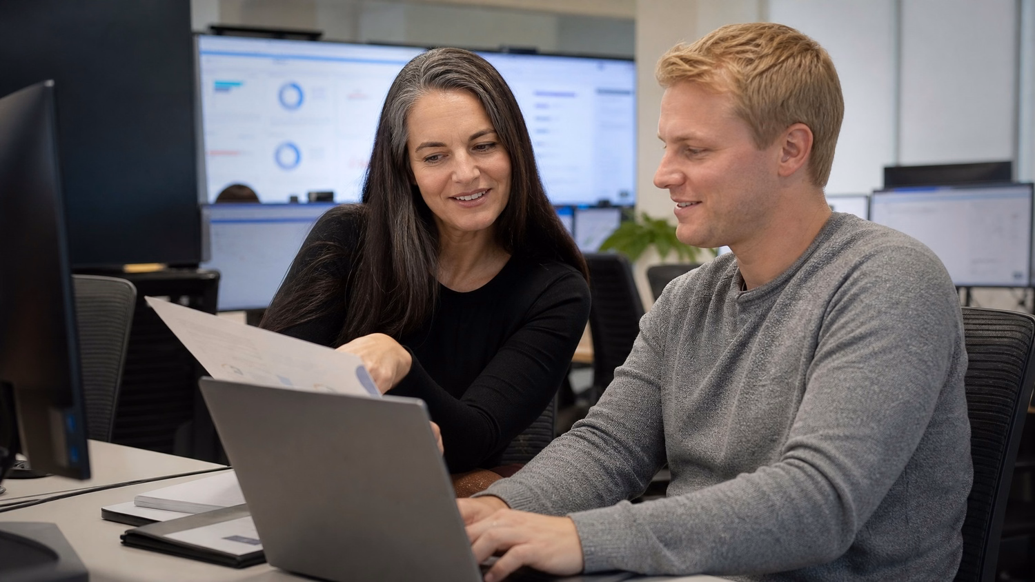 Woman shows document to man taking notes on laptop