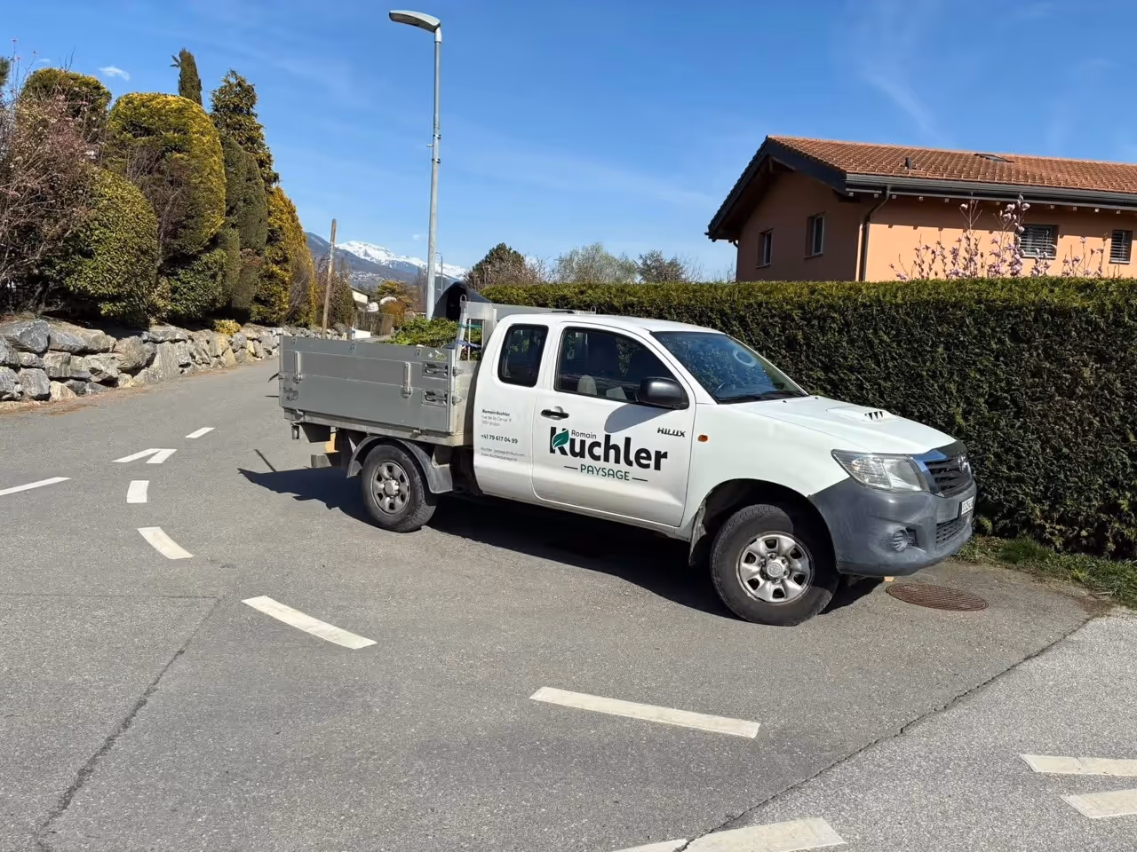Camionnette blanche avec logo d'aménagement paysager garée sur une route de quartier ensoleillée.