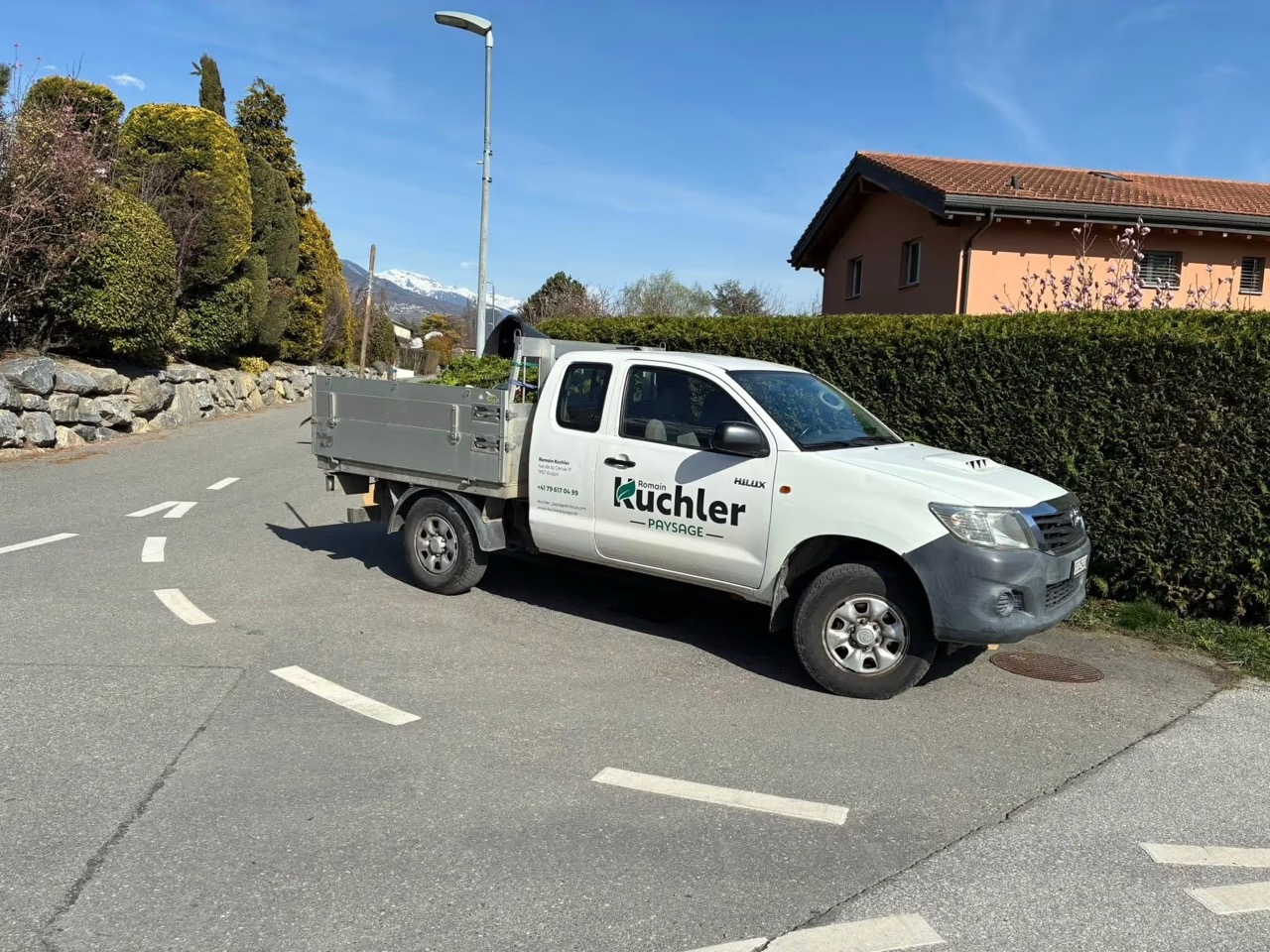 Camionnette blanche avec logo d'aménagement paysager garée sur une route de quartier ensoleillée.