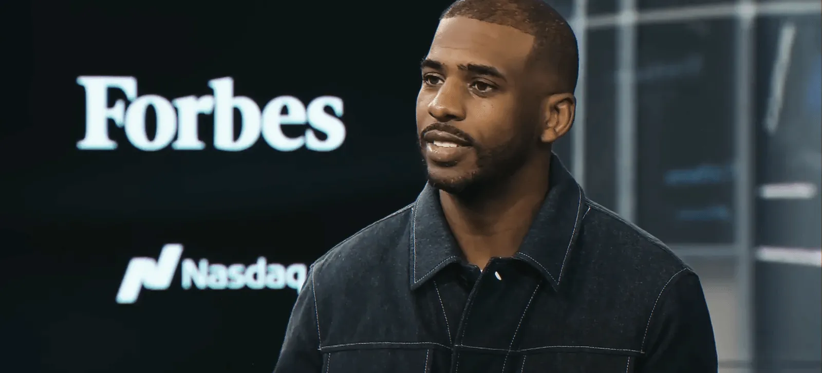 Chris Paul in a dark jacket, speaking in front of a screen displaying "Forbes" and "Nasdaq" logos.