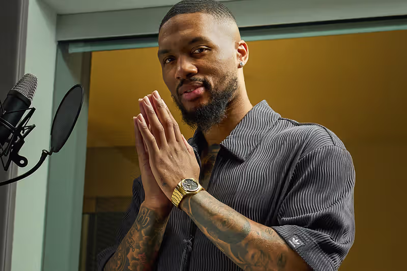Damian Lillard with hands clasped, wearing a gold watch, next to a microphone in a recording studio.