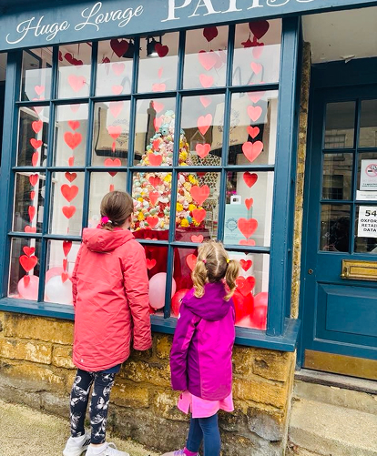 Two young girls in bright coats, on Burford high street, looking at confectionary shop window. 