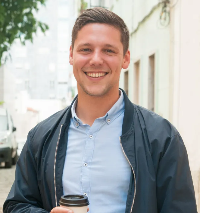 Smiling young man in a blue shirt and jacket holding a coffee cup outdoors on a city street.