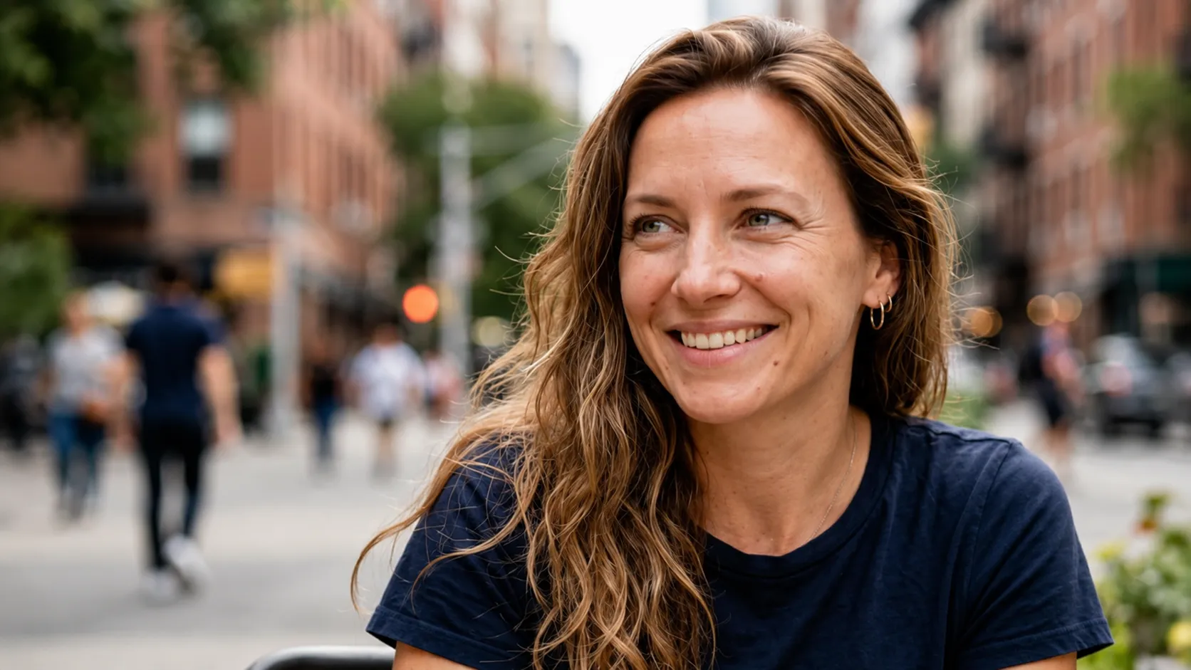 Smiling woman with wavy hair wearing a navy shirt sitting outdoors in a city street setting.