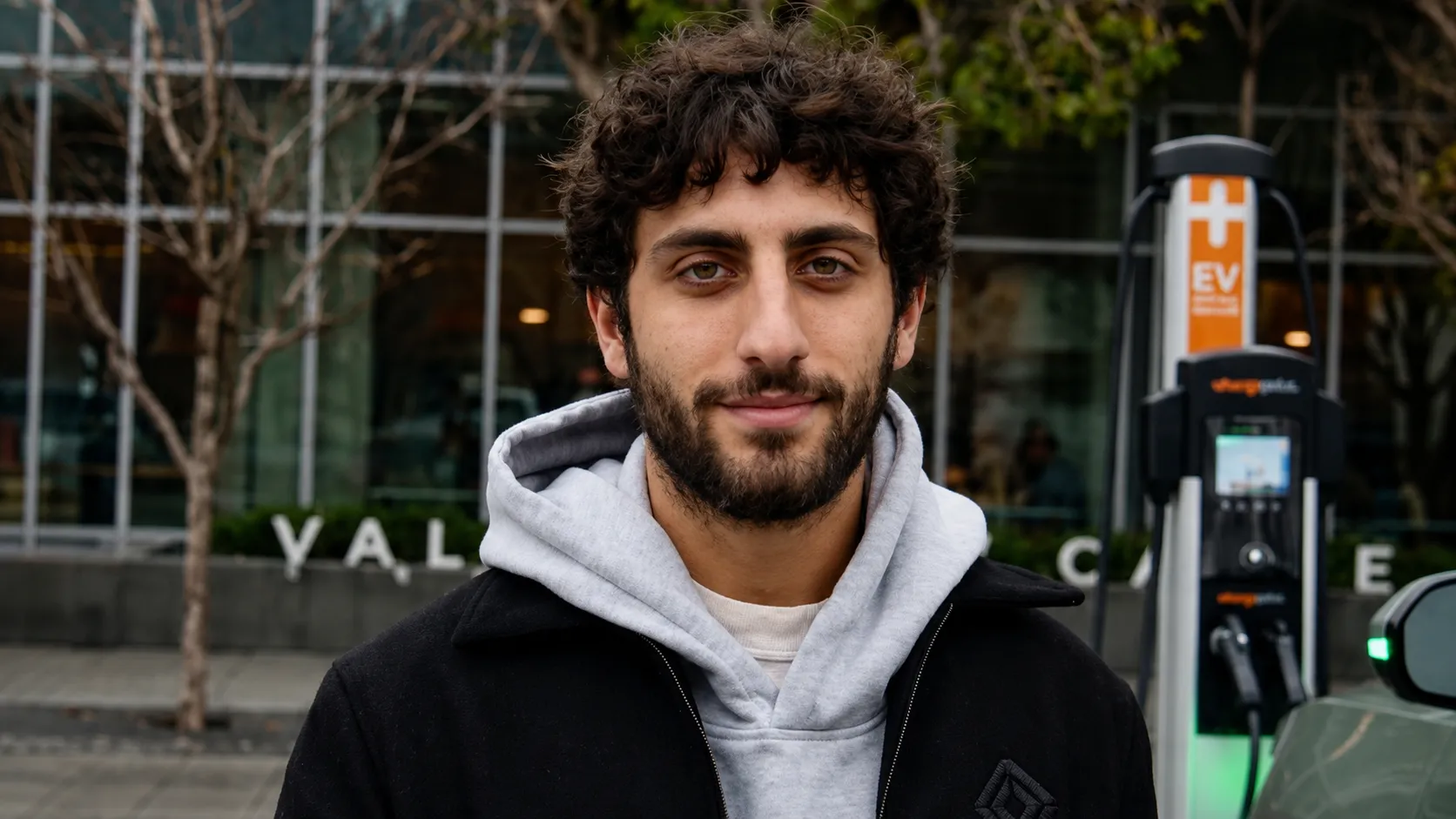 Man with curly hair and beard wearing a gray hoodie and black jacket by an EV charging station.