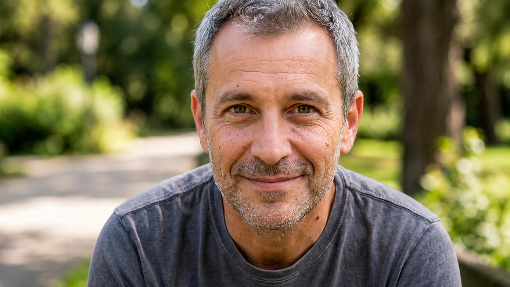 Smiling middle-aged man with gray hair and stubble wearing a gray shirt sitting outdoors.