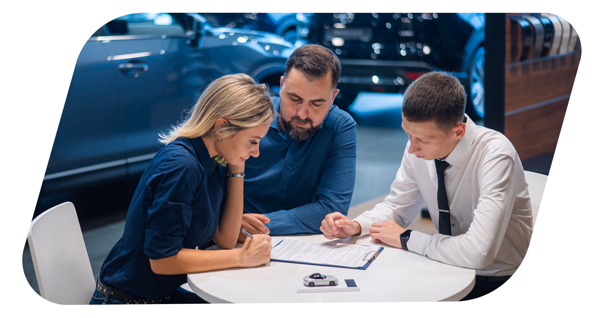 Three people at a car dealership table reviewing and signing documents with cars in the background.