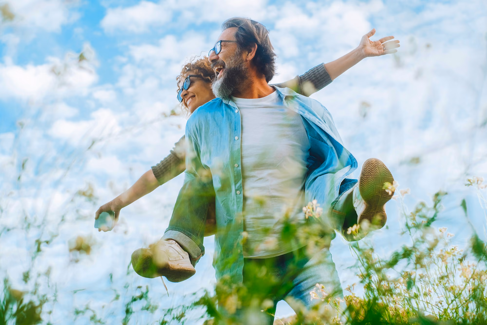 Man giving piggyback ride to woman outdoors in a field under a blue sky with clouds.