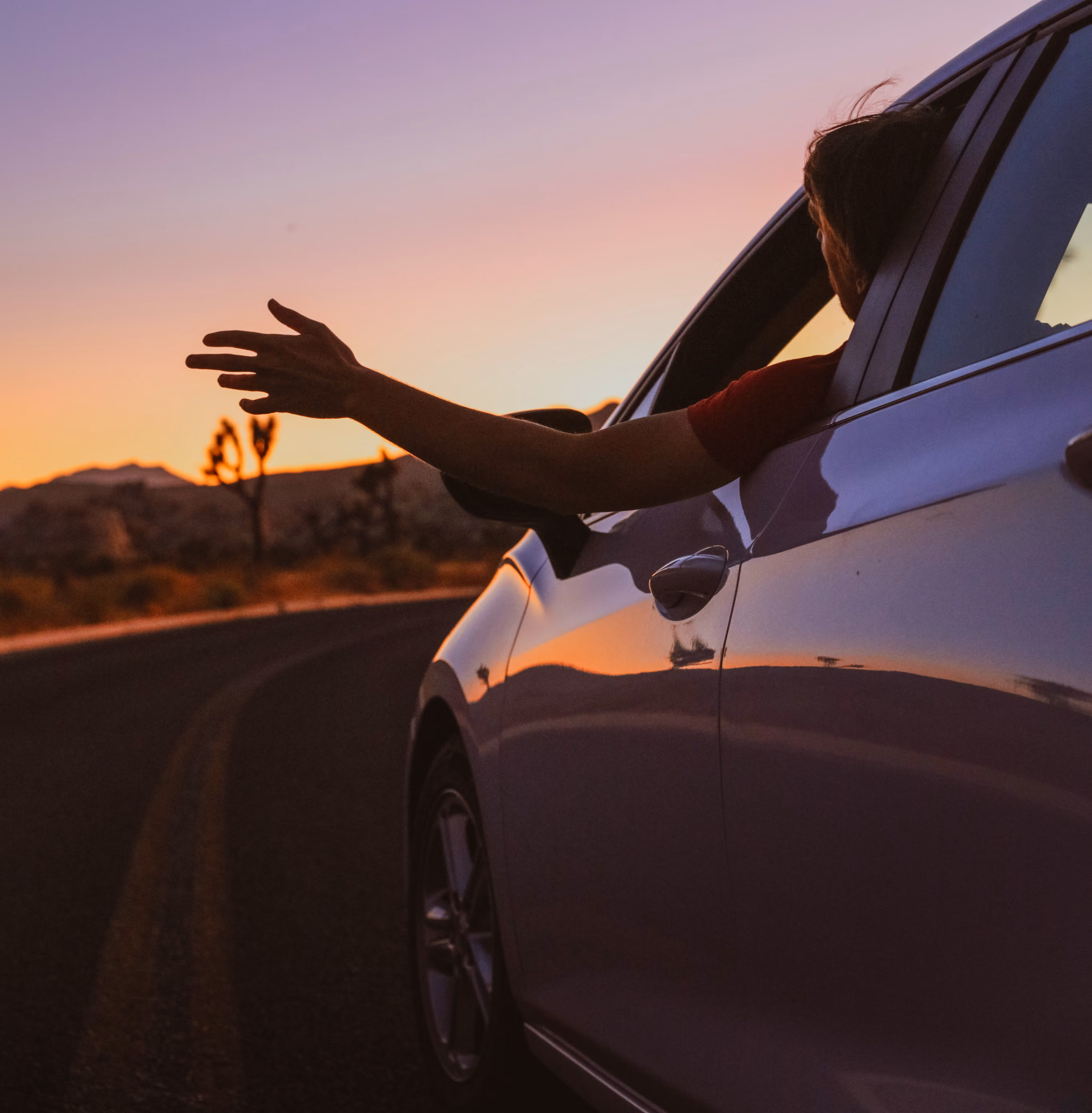 Person in red shirt extends arm out of car window at sunset on a desert road.
