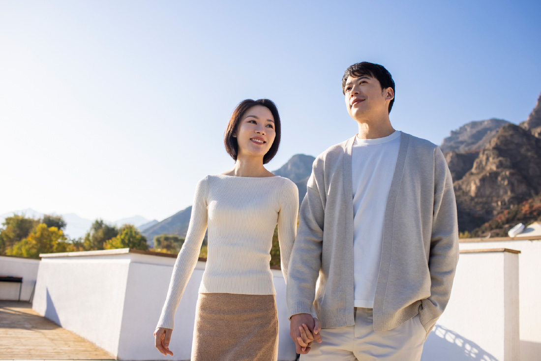 Couple holding hands on a sunny terrace, looking ahead with mountains behind them