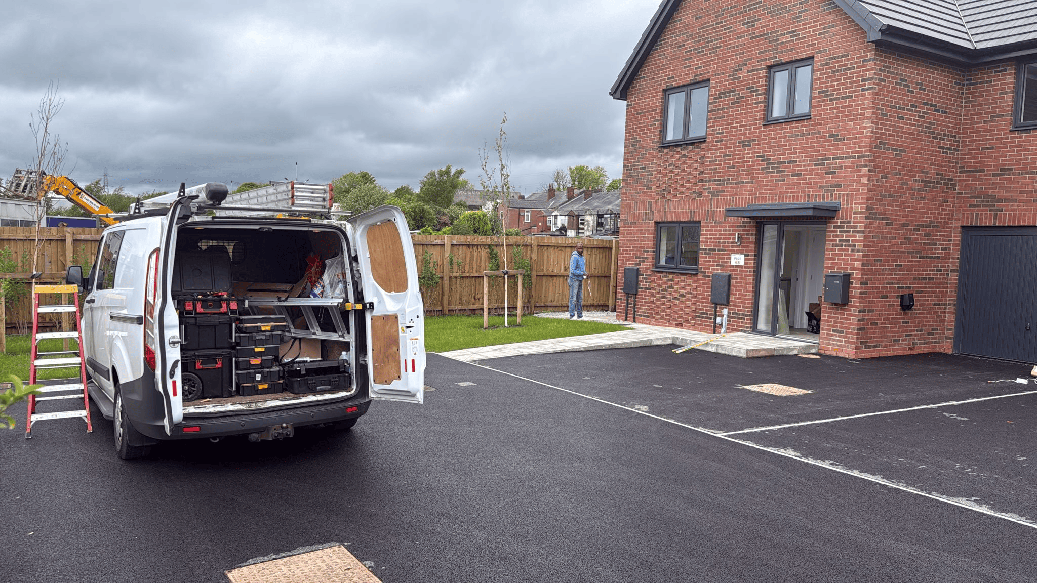 Worker standing near a brick house with an open van filled with toolboxes in a paved driveway.