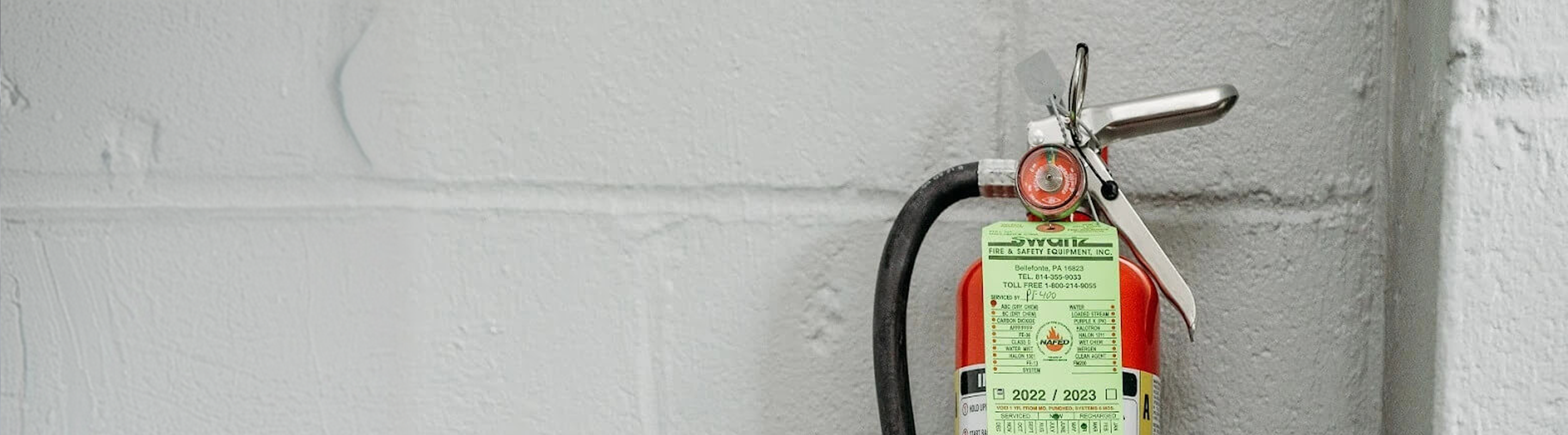 Red fire extinguisher mounted on a white brick wall with inspection tags from 2022 and 2023.