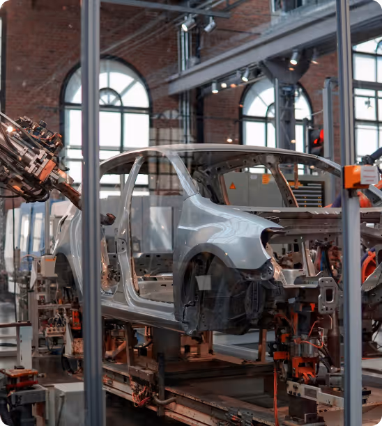 Automobile frame on assembly line with robotic arm welding inside factory with arched windows.