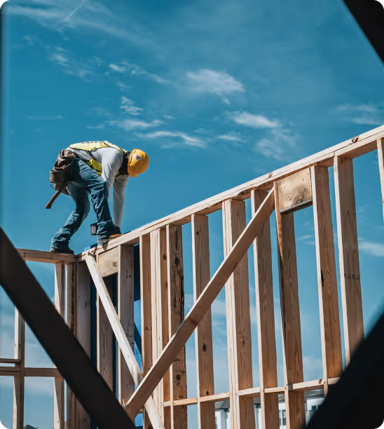 Construction worker in safety gear working on wooden frame structure under a blue sky.