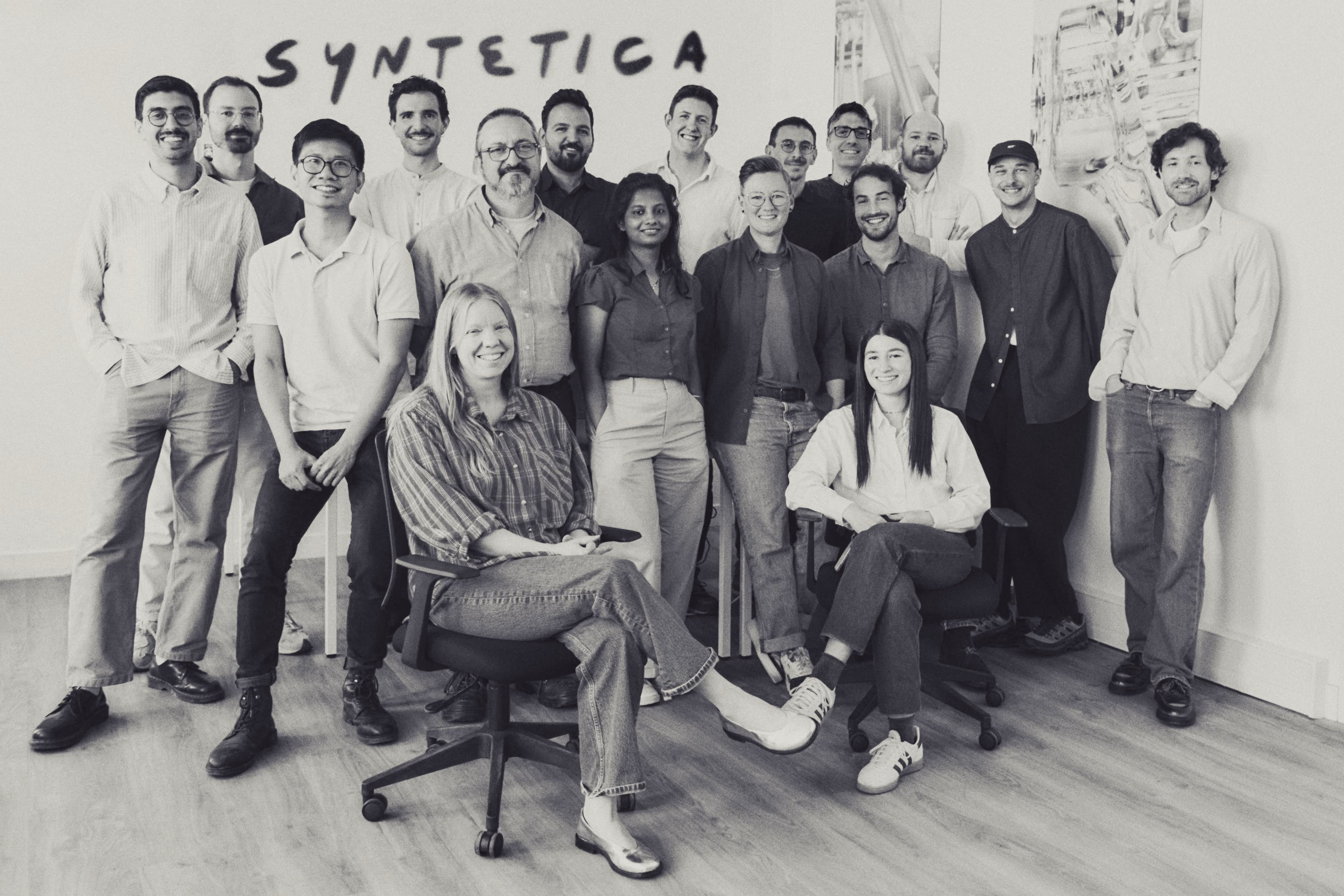 Group of 16 people posing indoors with SYNTHETICA sign on wall behind them.