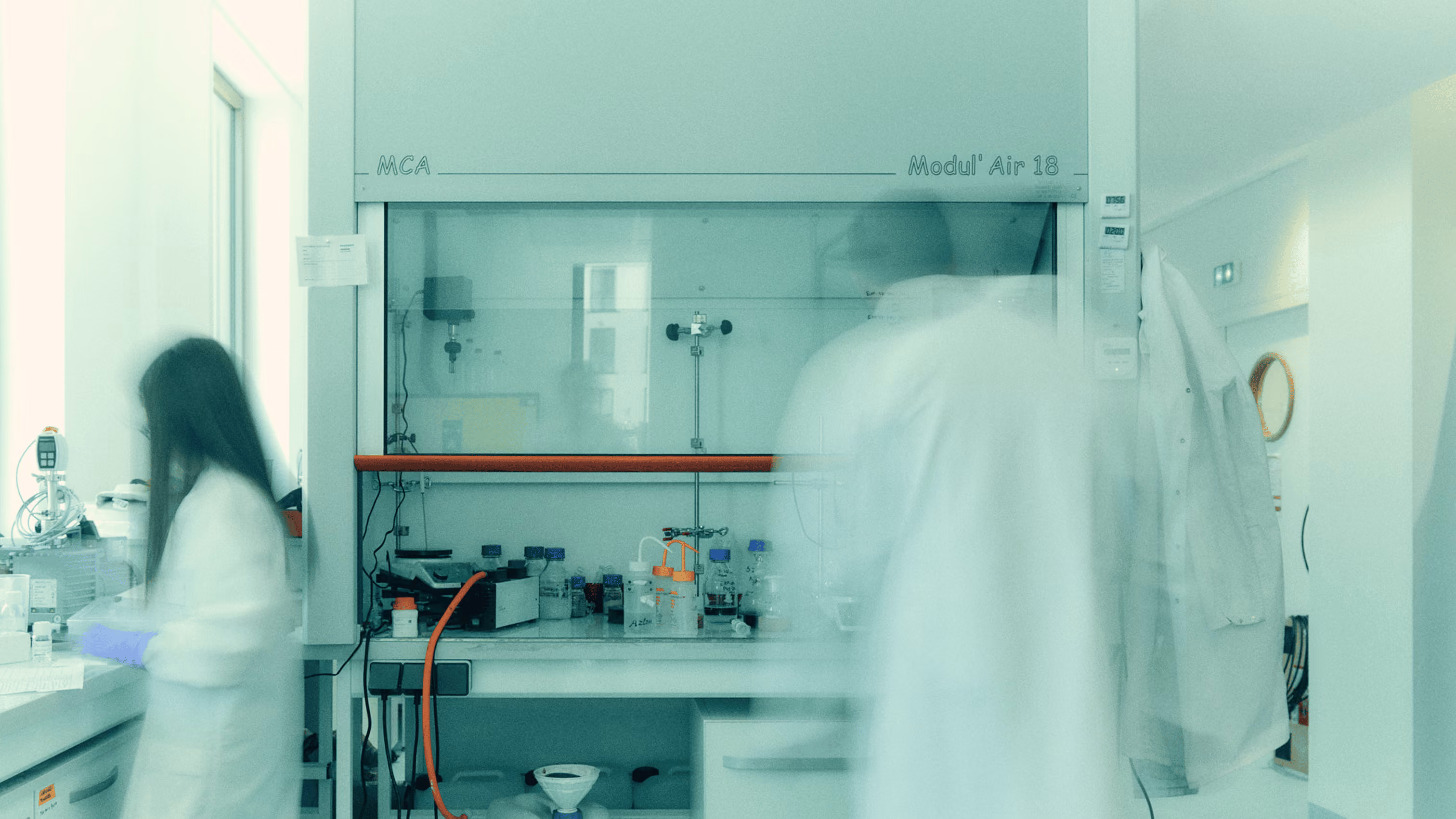 Two scientists in white lab coats working in a lab with chemical containers and equipment.