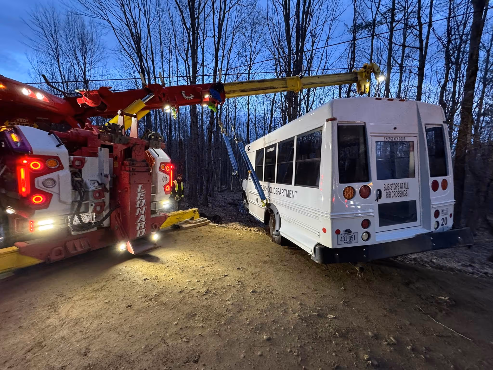 Tow truck lifting a school bus in a wooded area at dusk.