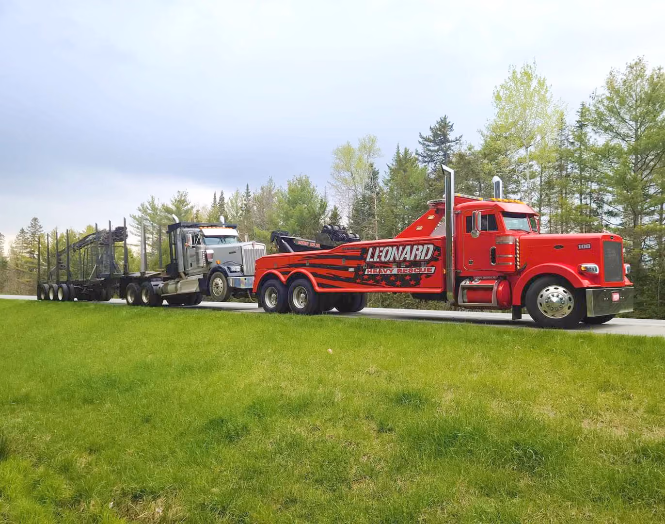 Red Leonard heavy rescue tow truck pulling a logging trailer on a roadside beside green grass and trees.