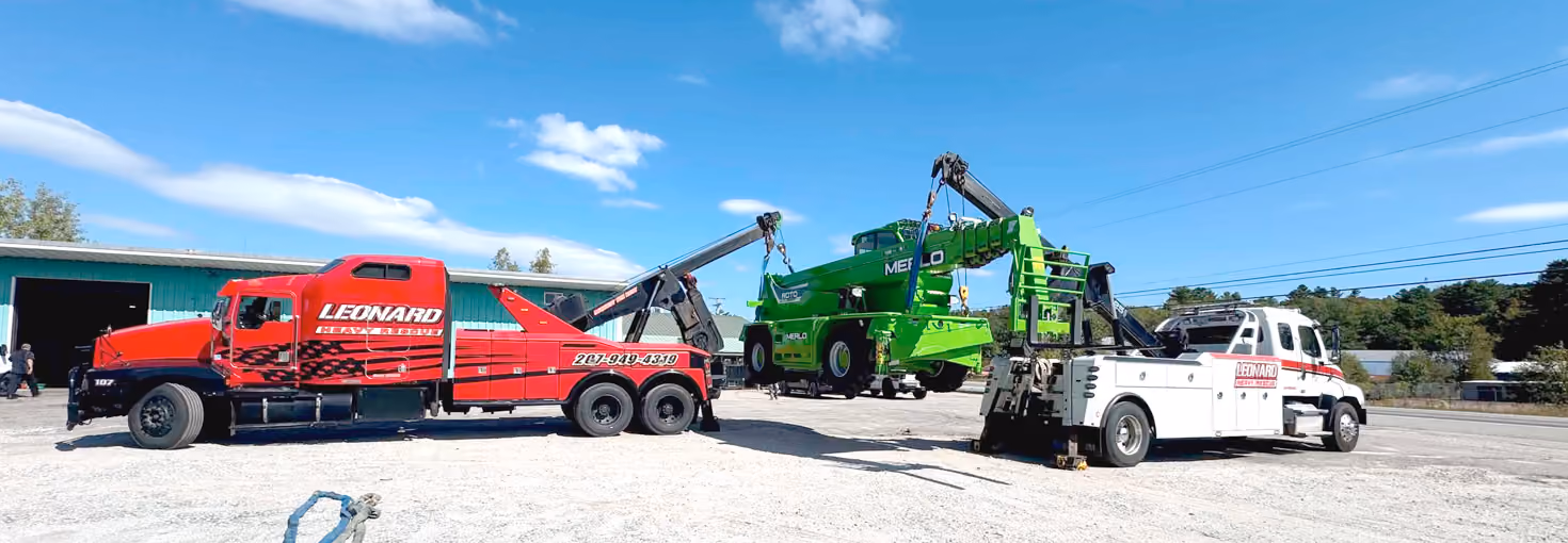 Two tow trucks lifting a green forklift in a parking lot under a blue sky.