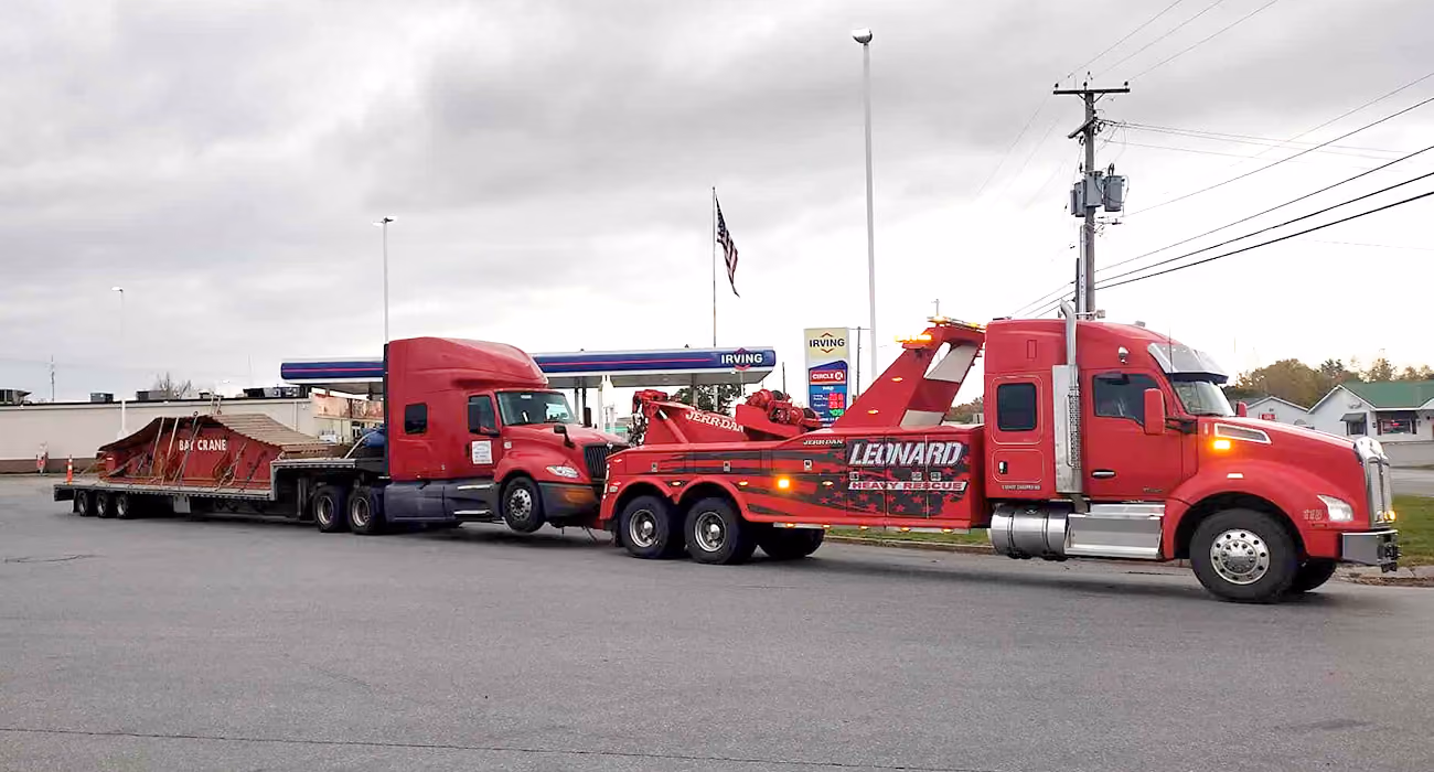Red heavy-duty tow truck hauling a large trailer on a roadside near a gas station.
