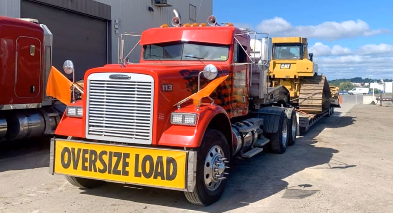 Red heavy-haul truck transporting a bulldozer on a flatbed trailer, with an oversize load sign.