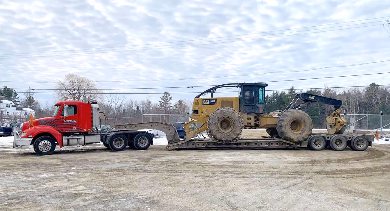 Red tow truck hauling a yellow CAT skidder on a lowboy trailer in a snowy lot.