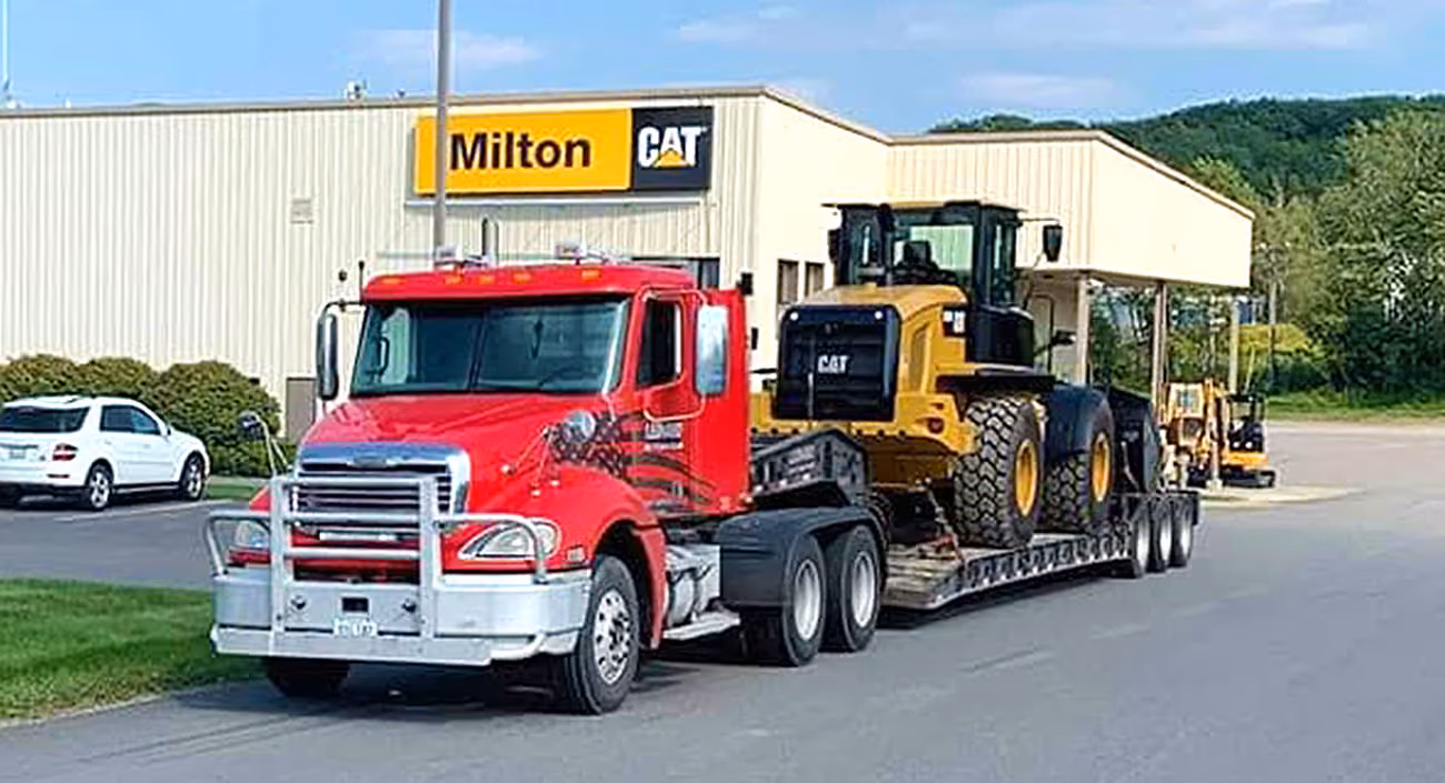 Red semi truck hauling a CAT bucket loader on a lowboy trailer outside a Milton CAT dealership.