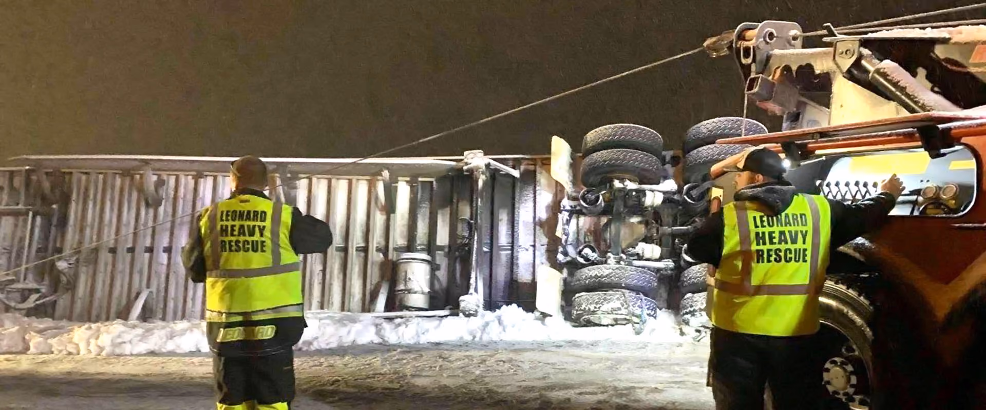 Two heavy rescue workers stand beside an overturned truck being recovered with a tow truck.