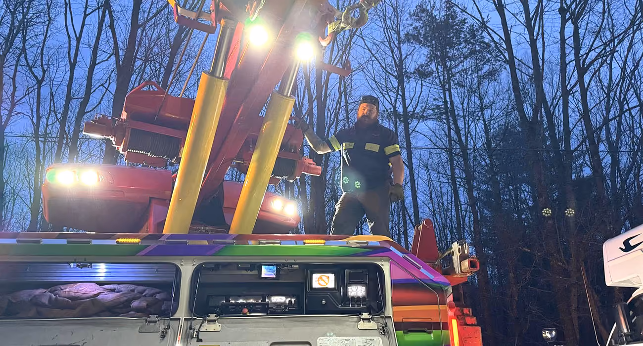 A worker in safety gear stands on a large red tow truck at dusk, illuminated by bright lights, with leafless trees and a dark blue sky in the background.