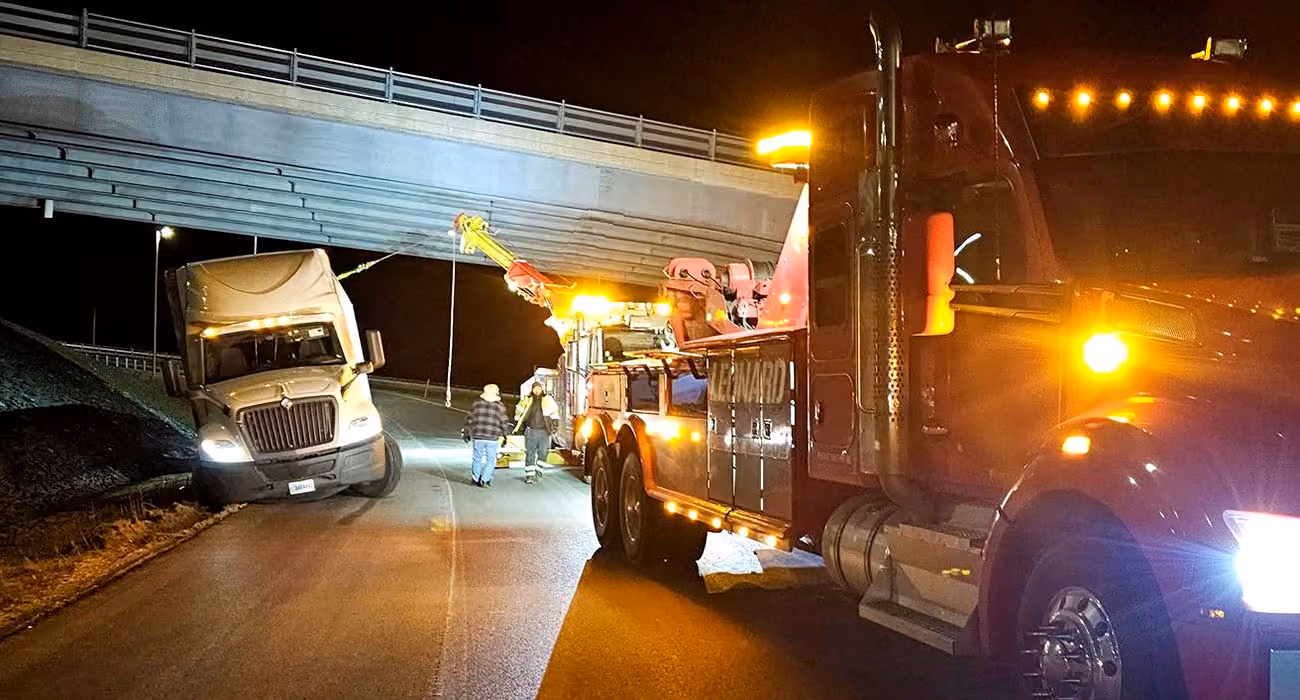 A tow truck with flashing lights assists a white semi-truck stuck under a highway overpass at night, while workers in reflective gear stand nearby.