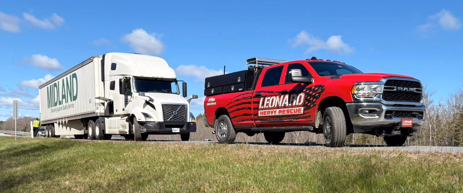 A white semi-truck with MIDLAND on the trailer is parked on the roadside behind a red pickup truck labeled LEONARD HEAVY RESCUE on a clear day with blue sky and clouds.
