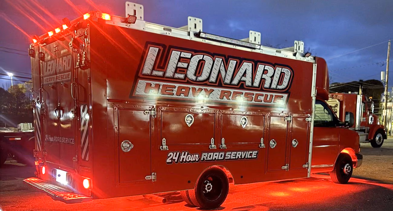 A bright red rescue truck labeled LEONARD HEAVY RESCUE and 24 Hour Road Service is parked at night with red lights illuminating the ground beneath it.