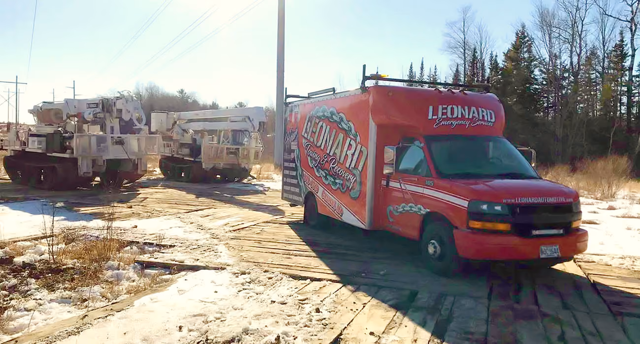 A red Leonard Emergency Services van is parked on a wooden plank road in a snowy, rural area, with utility trucks and trees in the background under a clear blue sky.
