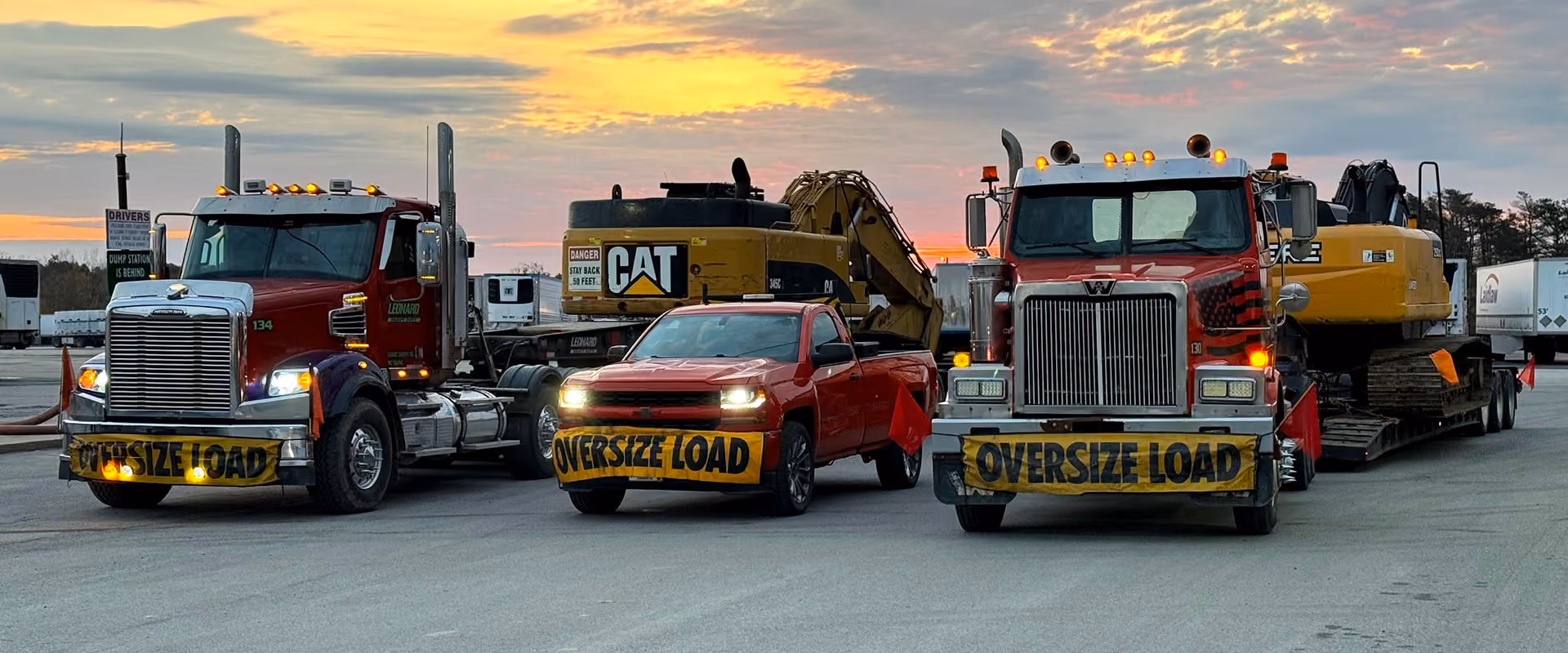 Three vehicles with "Oversize Load" banners. Two semi-trucks and a red SUV, parked in front of large construction equipment at sunset.