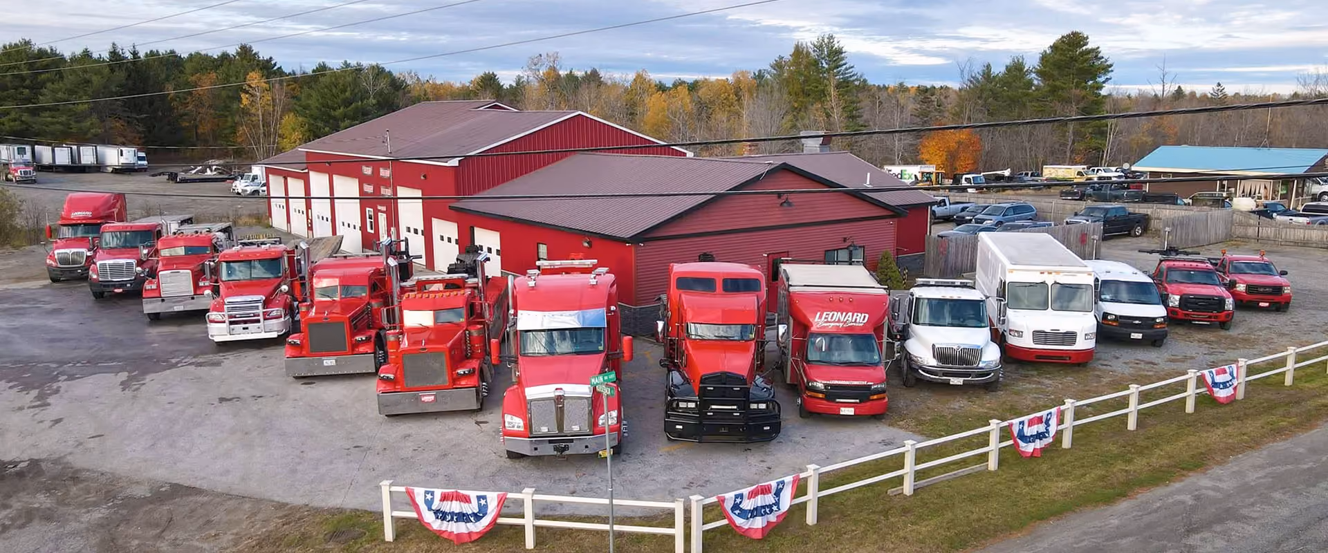 A row of red trucks, tow trucks, and vans parked in front of a large red garage building with white trim, surrounded by trees and a white fence.