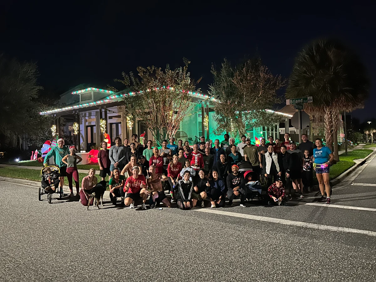 Large group of people smiling on street at night in front of house decorated with Christmas lights.