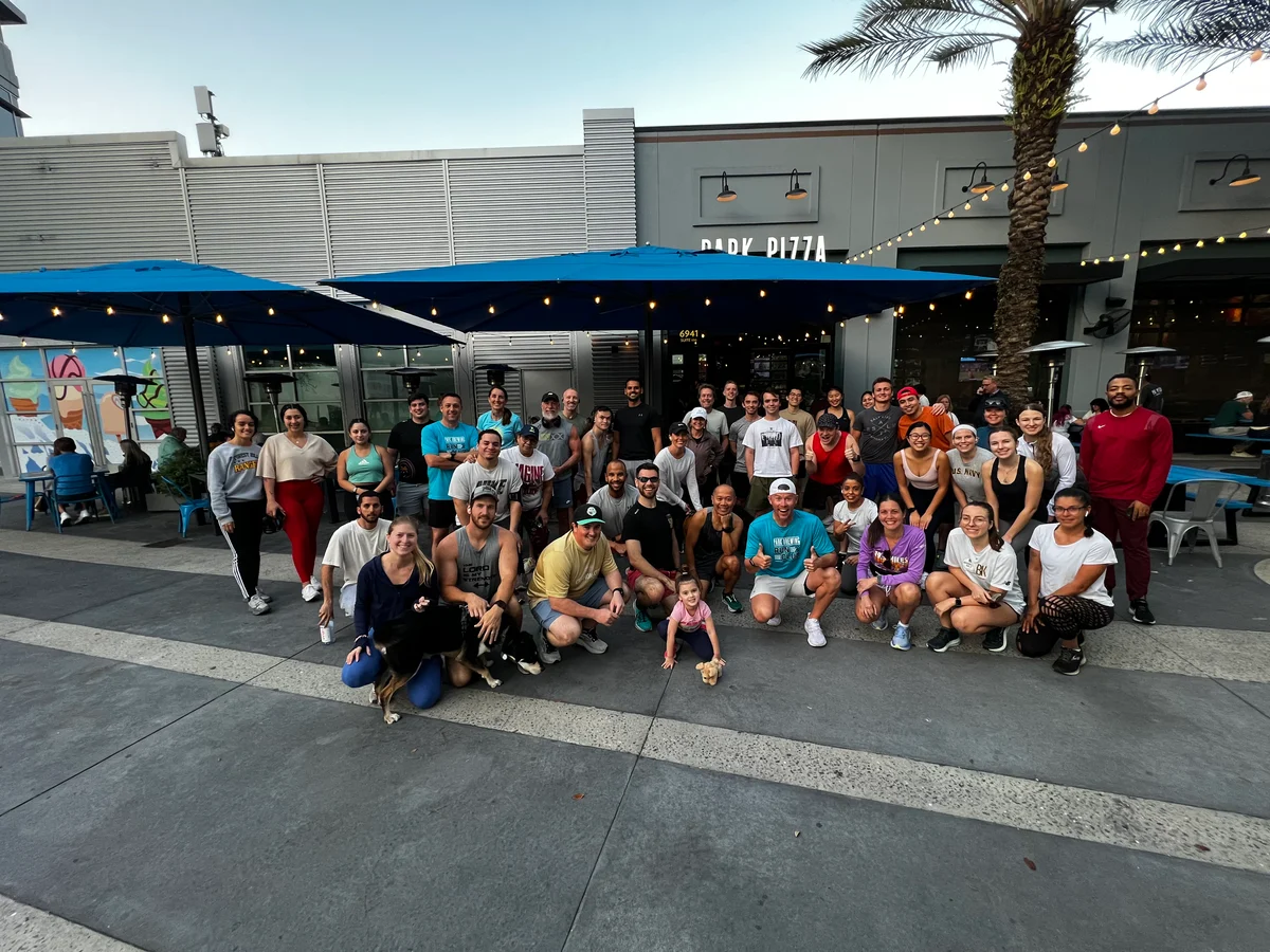 Large group of people posing outdoors in front of a restaurant with blue umbrellas and palm trees.