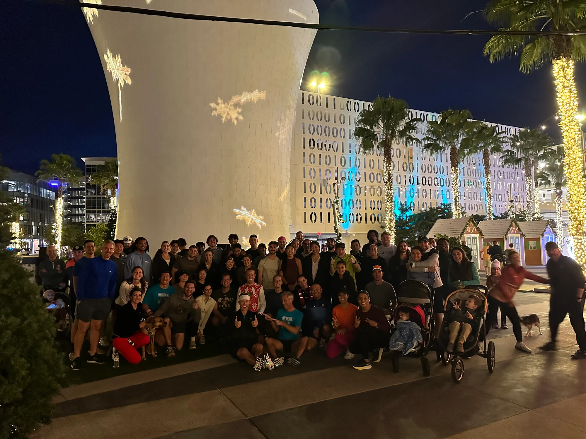 Large group of people posing outdoors at night with festive lights and snowflake projections in background.