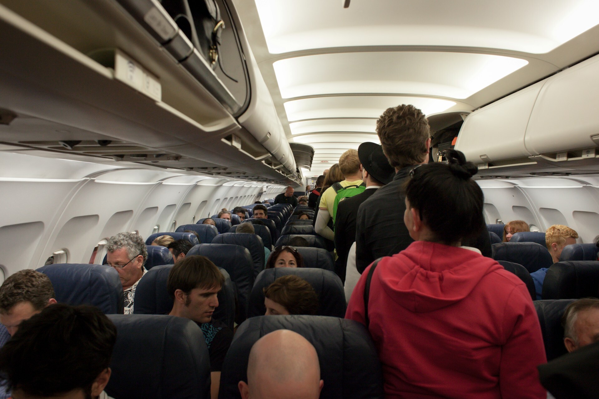 Photo taken in the inside of a plane looking down the walkway. Passengers are taking their seat as soon are sitting down and some are walking to their seat.