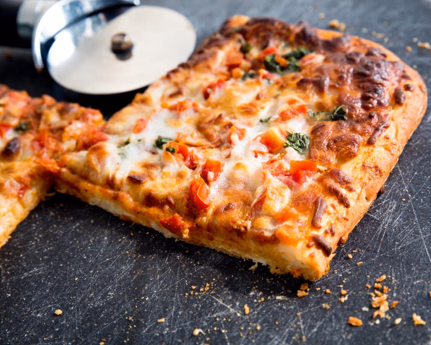 Square slice of cheesy pizza with tomatoes and spinach on a dark surface with a pizza cutter.