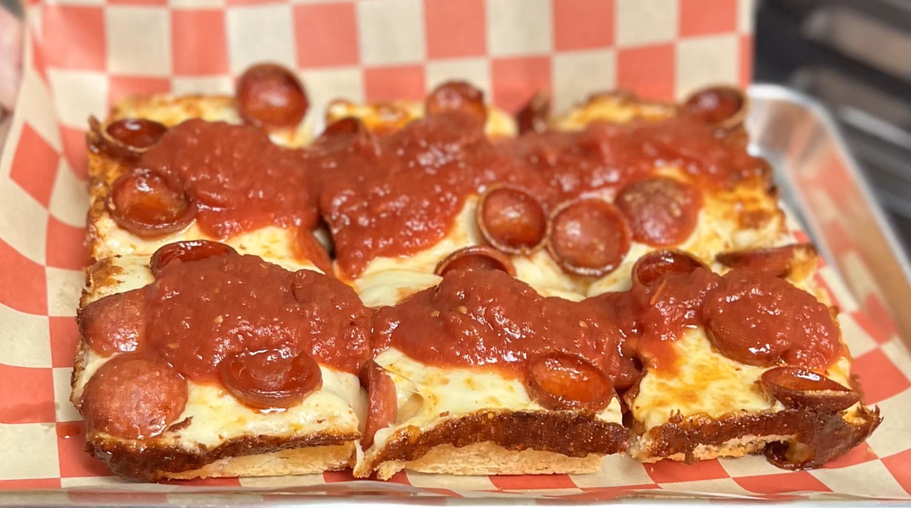 Square pepperoni pizza slices topped with marinara sauce on a red and white checkered paper.