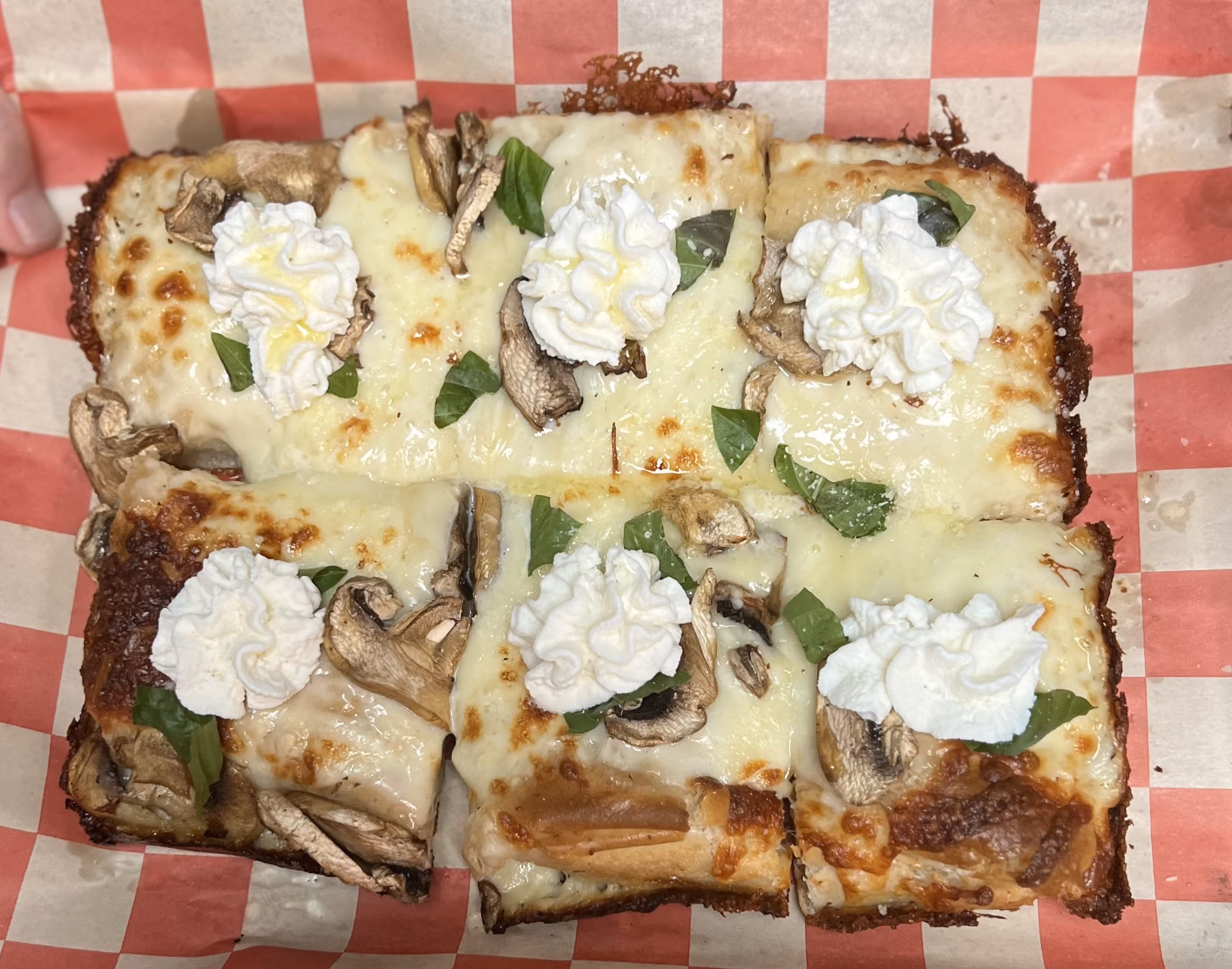 Square pizza slices with melted cheese, mushrooms, basil, and dollops of whipped cream on red and white paper.
