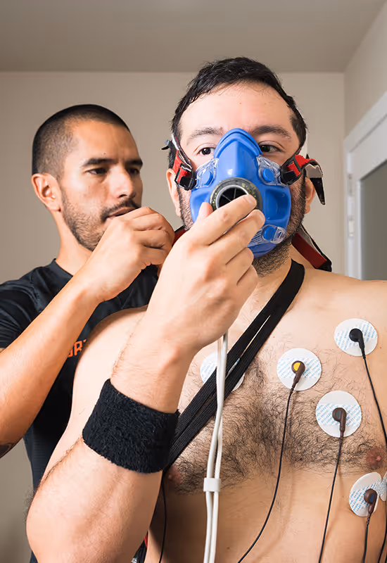 Hombre sin camisa usa máscara azul con electrodos en el pecho mientras otro ajusta la máscara.