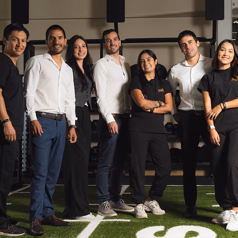 Grupo de siete personas sonrientes posando en un gimnasio sobre césped artificial.