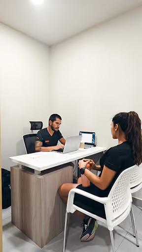 Hombre y mujer en consulta médica, él en escritorio con computadora, ella sentada frente a él.