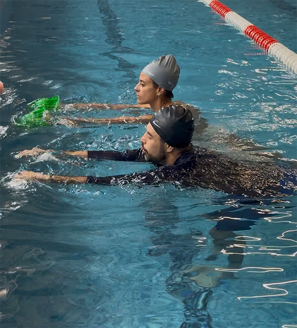 Dos personas con gorros de natación practican en una piscina, extendiendo brazos hacia adelante en el agua.